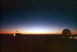 Both top and bottom of the image are black. In the middle of the image is the last sliver of the sun on the horizon. The edges of the horizon are blue and orange. On the left there is a lit-up triangle structure and on the right is a circle structure