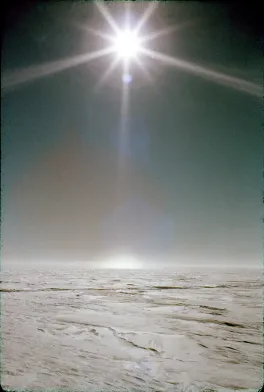 The sun is at the top of the image with halos around the sun and a bright spot on the ground below. The ground is covered with hard, flat snow.