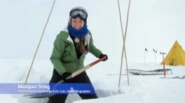 A white woman in snow gear with a shovel. She's standing in a shallow pit carved out of the surrounding snow. Behind her is a tent and a sled.