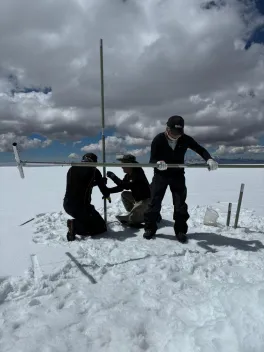 Three people drill into snow on a glacier under cloudy skies
