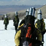 Many people with silver cylinders in their backpacks walking toward a dirt mountain in a distance on snow covered ground. 