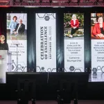 President Kristina Johnson on stage at a podium speaking with 4 hall length banners hanging of award winner images and text in the background at a distance