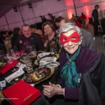 Ellen Mosley Thompson wearing a red eye mask smiling at the camera wearing a burgundy sweater and teal neck scarf with hands clutched together with short hair seated at a banquet table with food and a small red box on the table