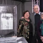 An ice sculpture on a table on the left and Georgie Shockey, Lonnie Thompson and Ellen Mosley Thompson standing next to it with Ellen clutching her arm with one hand while Lonnie is standing in the middle with his hands on both women's shoulders and Georgie resting right hand on the table to her right.