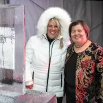 Andrea in white puff jacket with Eskimo hat standing next to an ice sculpture on a table to her right and Georgie Shockey to her right in a red black and gold embroidered jacket with black top under.