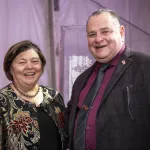 Georgie Shockey on the left posing indoor with her partner John with lilac color background walls. 
