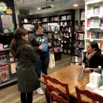 A person seated at a table in a bookstore speaking to two others.