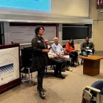 A person at the podium, while someone is speaking and a panel of four is seated next to the podium in the auditorium.