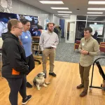 Three people and a dog listening to a presenter of a poster in an open space.