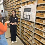 Two people discussing a poster in a repository with shelves of boxes.