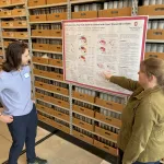 Two people discussing a poster in a repository with shelves of boxes.