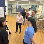 Three people listening to a presenter of a poster in an open space.