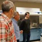 People in a laboratory setting, listening to a  person speaking.