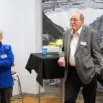Three individuals conversing at an event, with black and white landscape photographs on the wall and a floral arrangement on a nearby table.