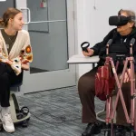 A person using virtual reality equipment is sitting across from another person in a casual indoor setting.