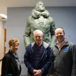 Three people standing beside a large statue of Admiral Byrd at a public facility.