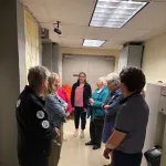 A group of people standing in a corridor, listening to a person in the center speaking.