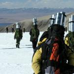 Many people with silver cylinders in their backpacks walking toward a dirt mountain in a distance on snow covered ground. 
