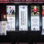 President Kristina Johnson on stage at a podium speaking with 4 hall length banners hanging of award winner images and text in the background at a distance
