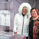 Andrea in white puff jacket with Eskimo hat standing next to an ice sculpture on a table to her right and Georgie Shockey to her right in a red black and gold embroidered jacket with black top under.