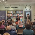Two people seated and one person at the podium talking with an audience in a book store.