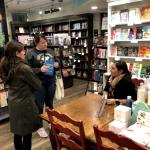 A person seated at a table in a bookstore speaking to two others.