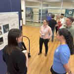 Three people listening to a presenter of a poster in an open space.