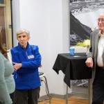 Three individuals conversing at an event, with black and white landscape photographs on the wall and a floral arrangement on a nearby table.