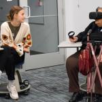 A person using virtual reality equipment is sitting across from another person in a casual indoor setting.